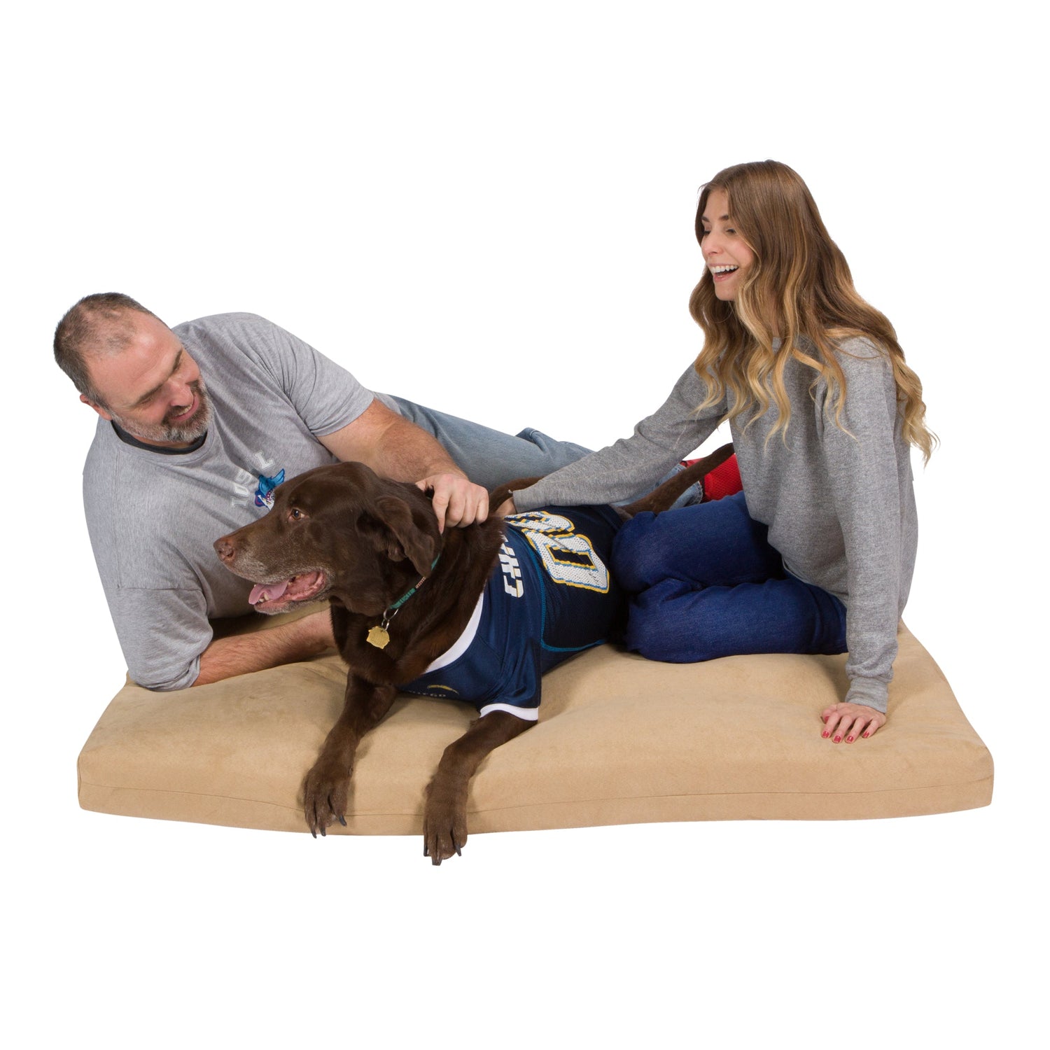 A man and woman smile and pet a large brown dog in a sports jersey while sitting on a beige Temperature Regulated Orthopedic Dog Bed. The cozy bed’s gel-infused memory foam ensures comfort, with a white background highlighting their joyful moment.